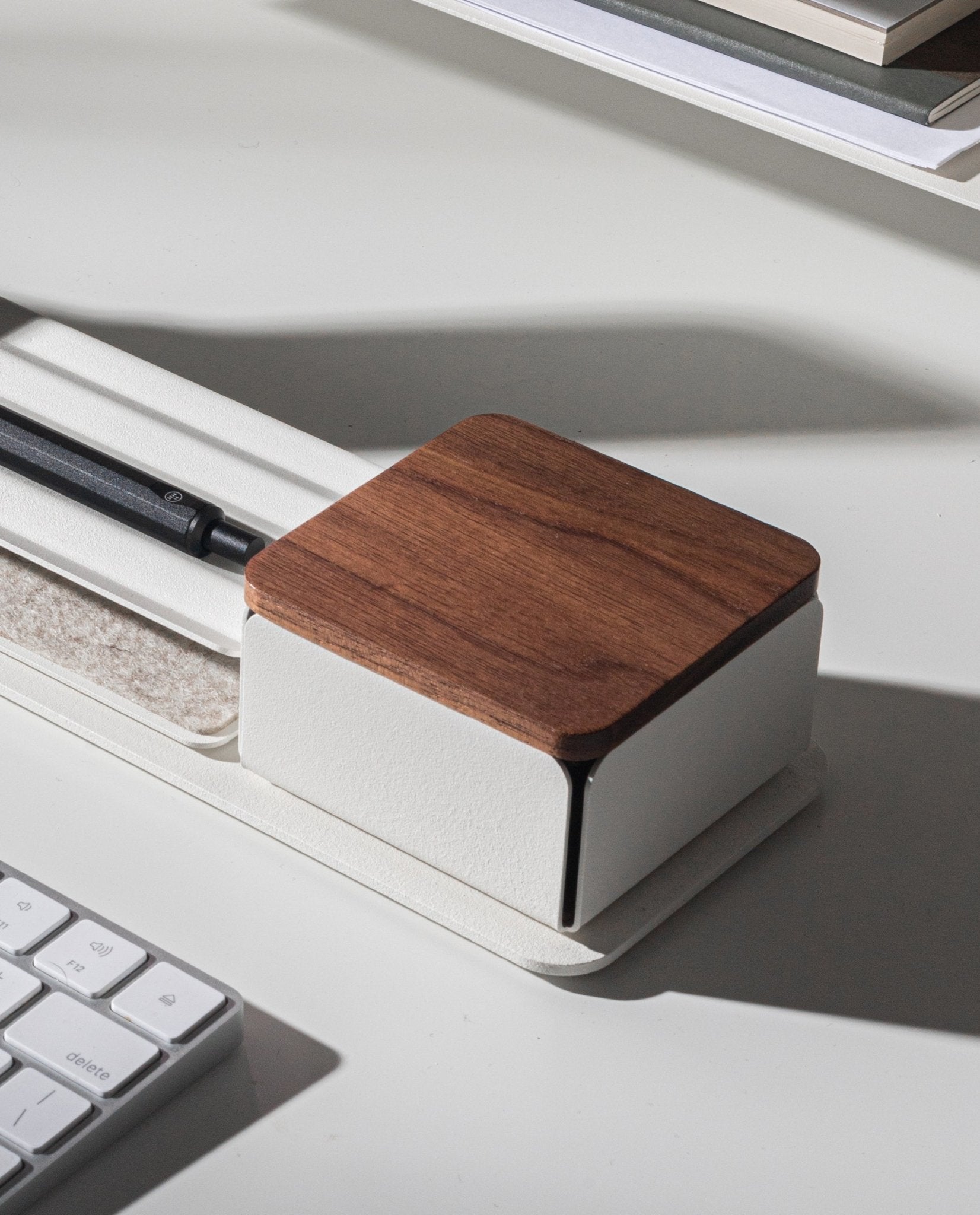 A Gather Solid Wood Lid (Walnut) organizer, featuring a black pen inside and a white base, sits on a white desk near a computer keyboard and stacked books.
