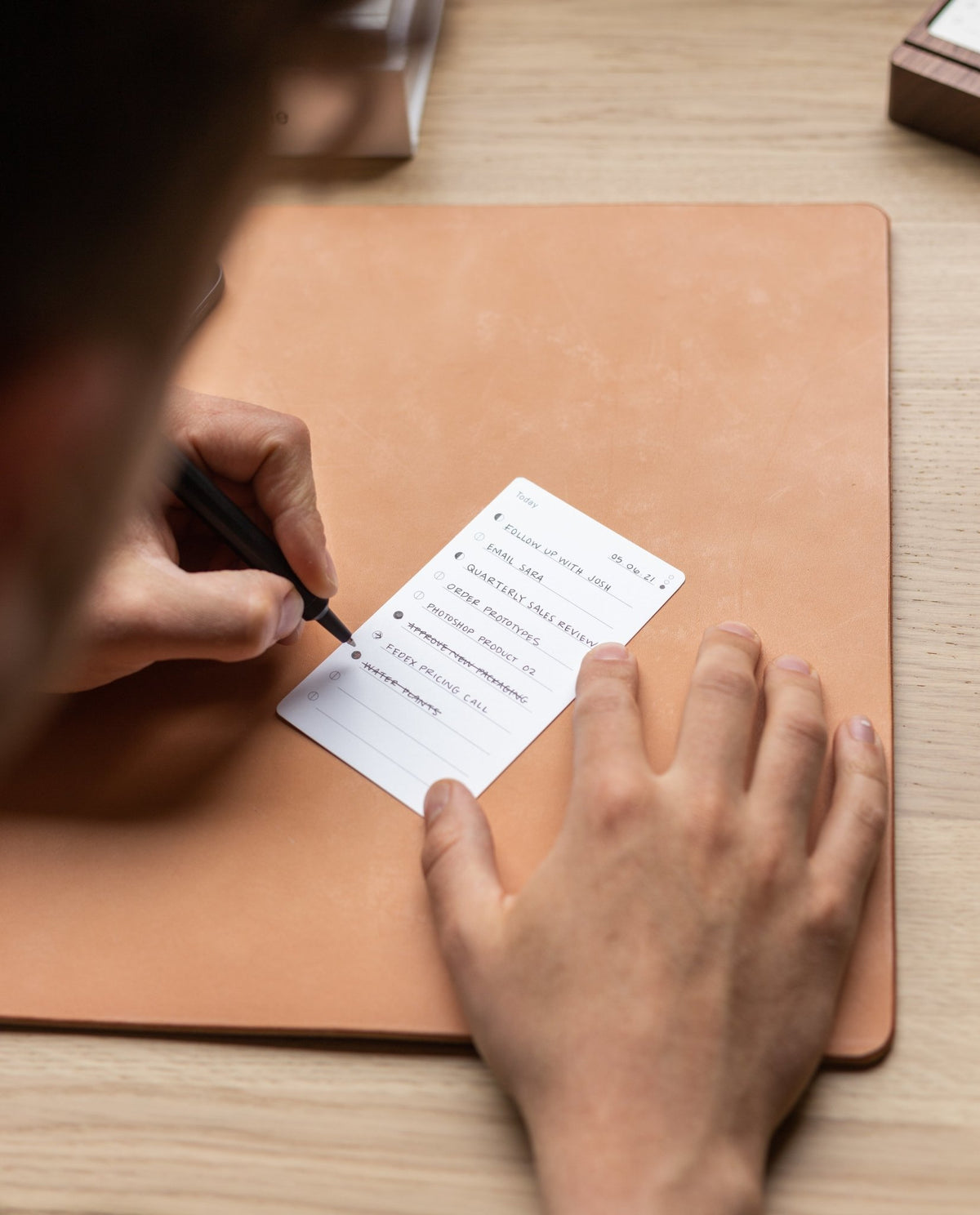 A person uses the Analog Premium Starter Kit (Walnut) as a task organizer, writing to-do items on its notepad with a black pen atop a tan desk mat to enhance personal productivity.