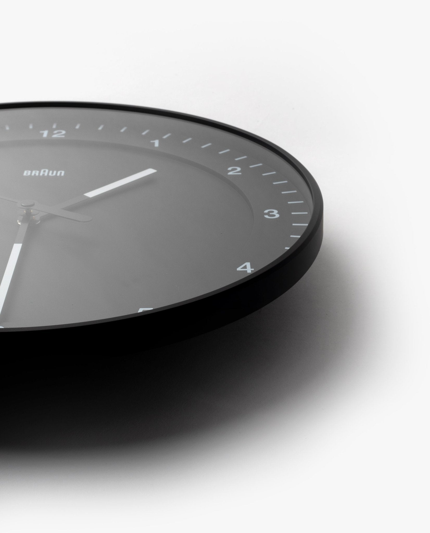 The Braun Wall Clock (Black) features minimalist white hour markers and hands, shown at an angle with only the lower right of the face visible against a white background. An easy-to-read clock, ideal for any modern space.