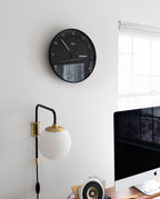 A modern, minimal office features the Braun Wall Clock (Black) displaying 12:14, a round wall lamp, part of a computer monitor, and a window letting in daylight.
