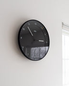 A Braun Wall Clock (Black) with white numbers and hands displays 10:10. Mounted on a white wall near a window, it’s easy to read in daylight streaming through.