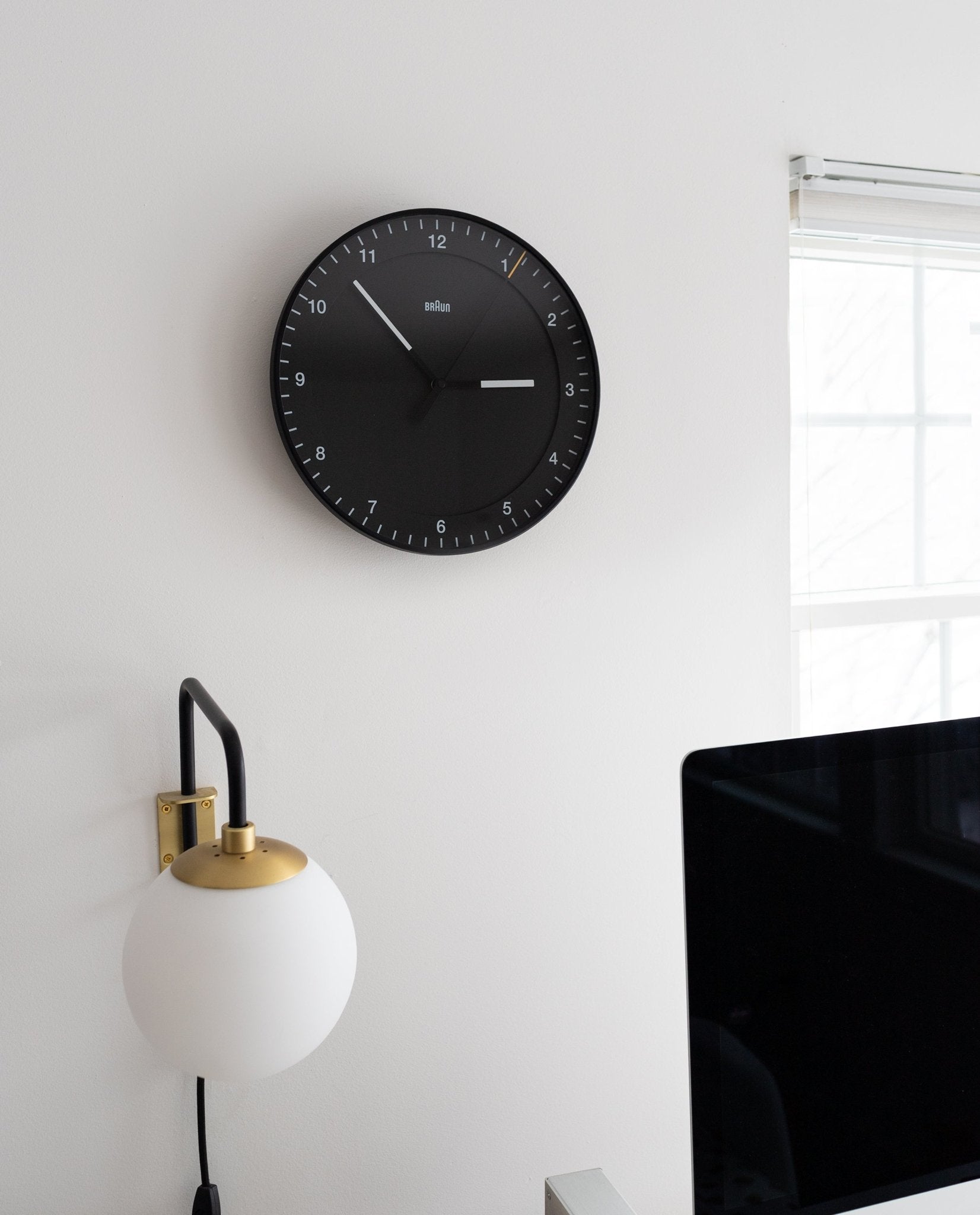 A Braun Wall Clock (Black) showing 10:08 hangs on a white wall above a globe-shaped lamp by a window. Its easy-to-read face and silent mechanism promote calm, with the edge of a computer monitor visible in the foreground.