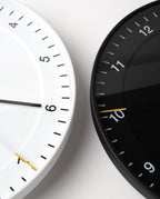 Two Braun Wall Clocks, one in white and one in black, are shown side by side, each featuring an easy-to-read dial and modern design. Both display the time as 6:50. The clocks are cropped to reveal only portions of their faces against a pale background, drawing attention to the Braun Wall Clock (White) and its silent sweeping second hand.