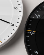 Two Braun Wall Clocks overlap on a light surface. The left clock, Braun Wall Clock (White), displays 5:30, while the right clock is black showing 10:45. Reminiscent of the classic design, both feature minimalist dials with easy-to-read faces and yellow silent sweeping second hands.