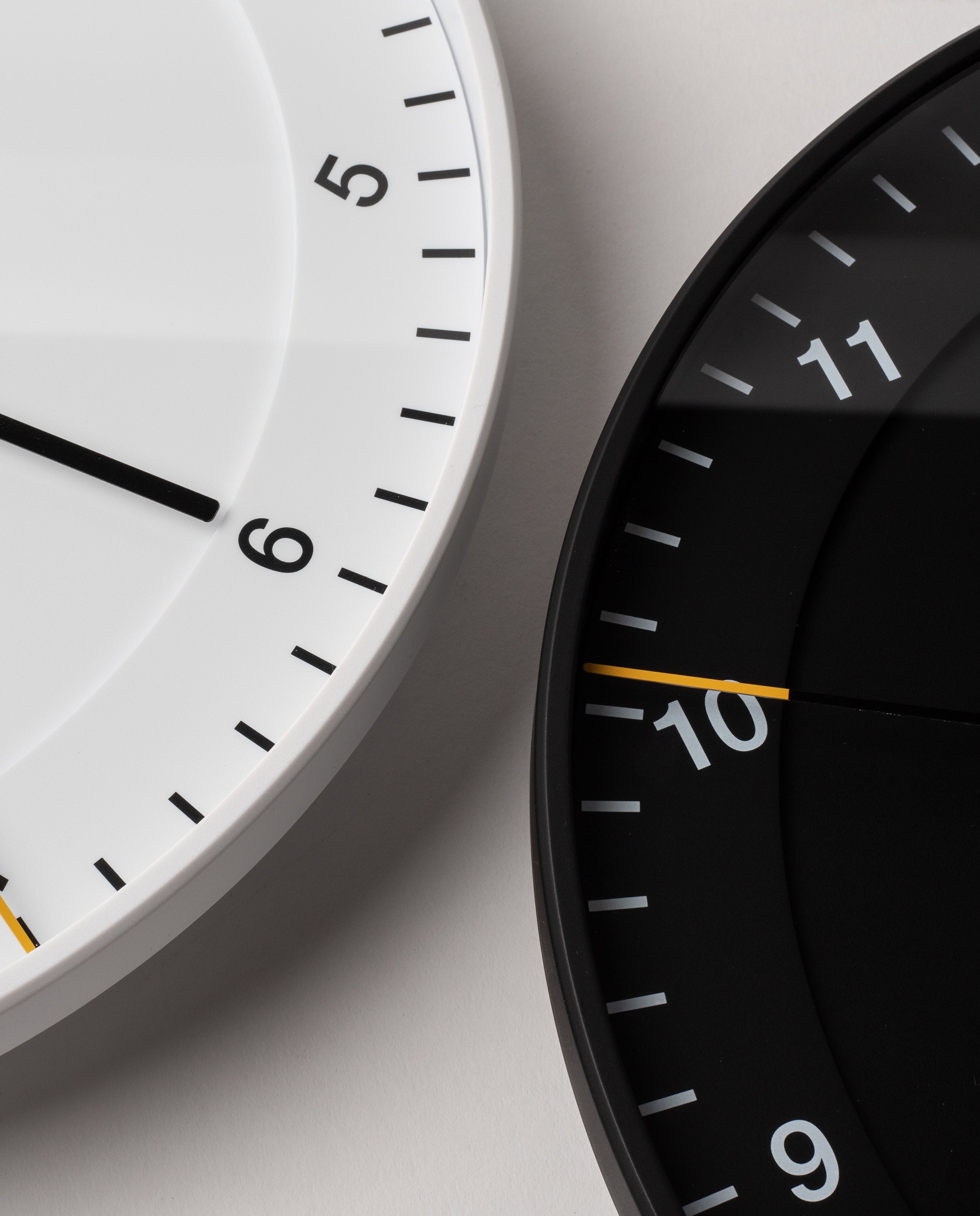 Two Braun Wall Clocks overlap on a light surface. The left clock, Braun Wall Clock (White), displays 5:30, while the right clock is black showing 10:45. Reminiscent of the classic design, both feature minimalist dials with easy-to-read faces and yellow silent sweeping second hands.