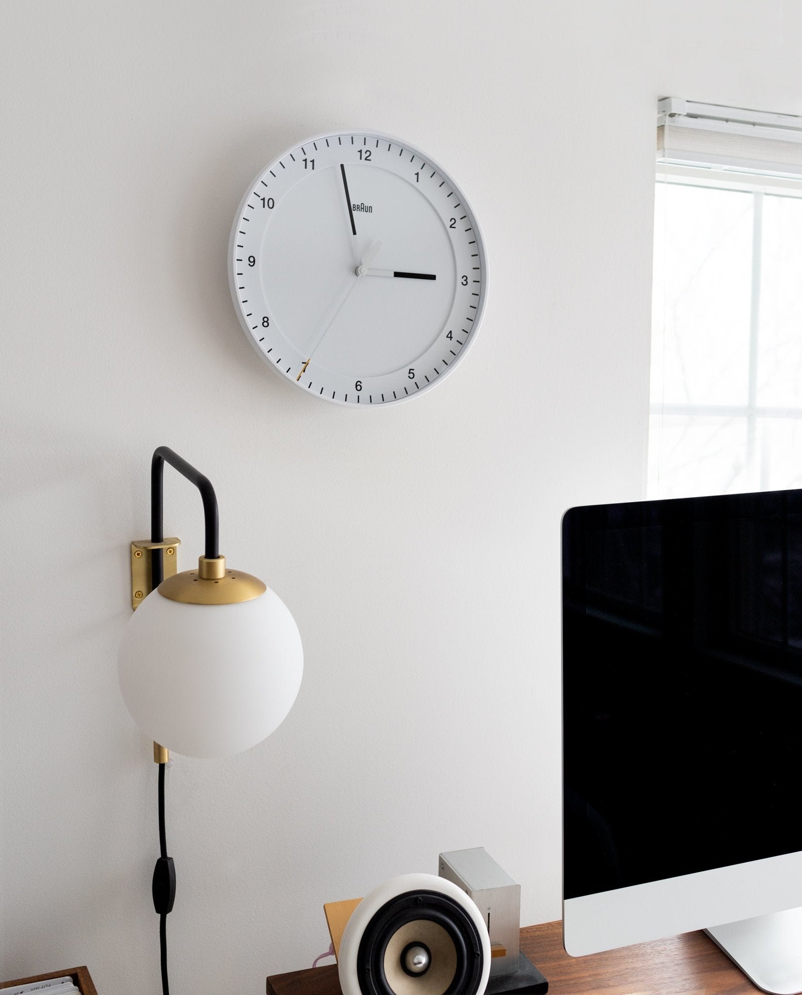 A Braun Wall Clock (White) with an easy-to-read dial and a silent sweeping second hand shows 3:38 above a globe wall lamp. A computer monitor, speaker, and small objects are arranged on a wooden desk by a window with white blinds.
