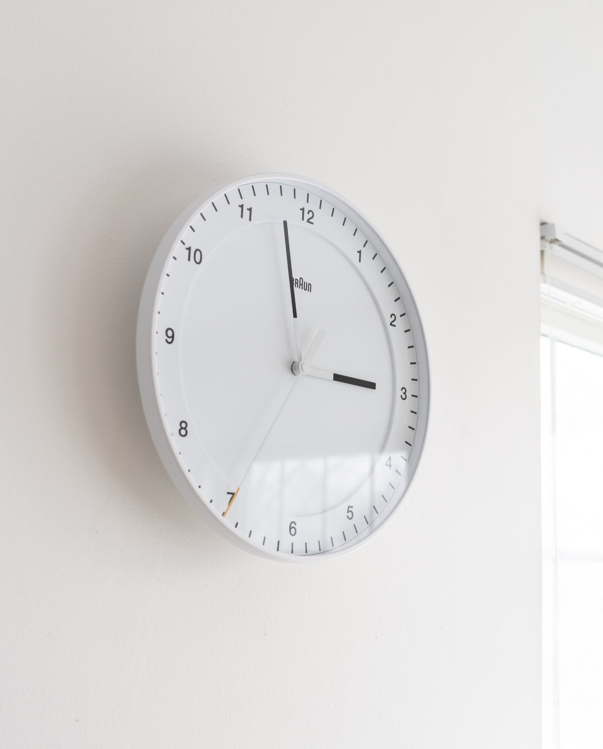 A Braun Wall Clock (White) displays the time as 2:56:35. The clock, which features an easy-to-read dial, is mounted on a white wall beside a window with blinds, illuminated by natural light streaming in.