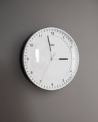 A Braun Wall Clock (White) with an easy-to-read dial features black hour and minute hands and a thin white silent sweeping second hand, set against a gray wall and displaying the time as 3:00:34.