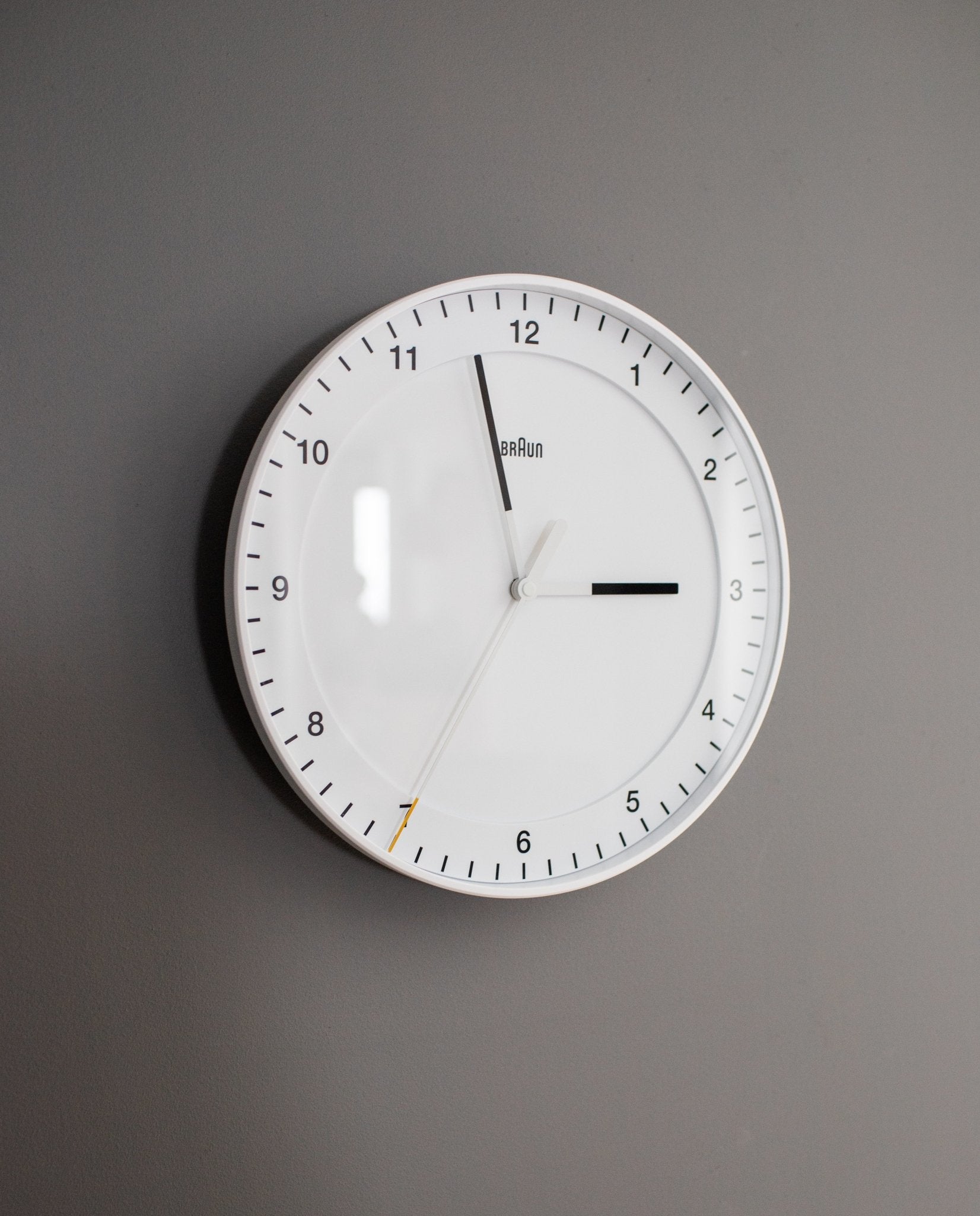 A Braun Wall Clock (White) with an easy-to-read dial features black hour and minute hands and a thin white silent sweeping second hand, set against a gray wall and displaying the time as 3:00:34.