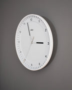 A Braun Wall Clock (White) with black hour and minute markers hangs on a gray wall, displaying the time 12:20:32. This clock showcases a minimalist design featuring thin black hands, a silent sweeping second hand, and an easy-to-read numberless dial.