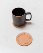 A Hasami Porcelain Mug (Matte Black) sits on a round tan coaster, with another nearby embossed with "ENJOY THE JOURNEY," all resting on a white marble surface.