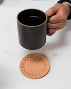 A hand holding a Hasami Porcelain Mug (Matte Black) above a round tan coaster embossed with "ENJOY THE JOURNEY," set on a white surface.
