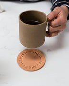 A hand holds the Hasami Porcelain Mug (Natural) above a round leather coaster embossed with "ENJOY THE JOURNEY," set on a white surface and evoking the timeless artistry of Japanese ceramics.