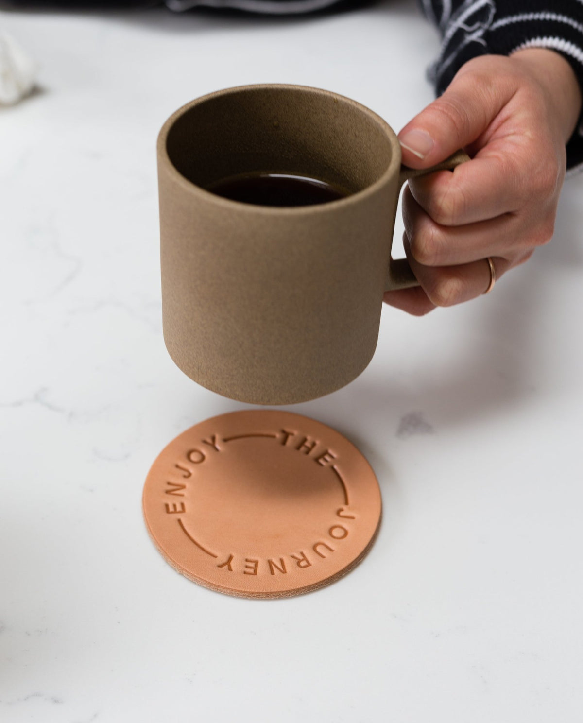 A hand holds the Hasami Porcelain Mug (Natural) above a round leather coaster embossed with "ENJOY THE JOURNEY," set on a white surface and evoking the timeless artistry of Japanese ceramics.