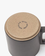 A close-up of an upside-down Hasami Porcelain Mug (Matte Black) shows its tan bottom stamped with "HASAMI PORCELAIN JAPAN," emphasizing its handmade quality. The mug’s handle is visible on the right side.