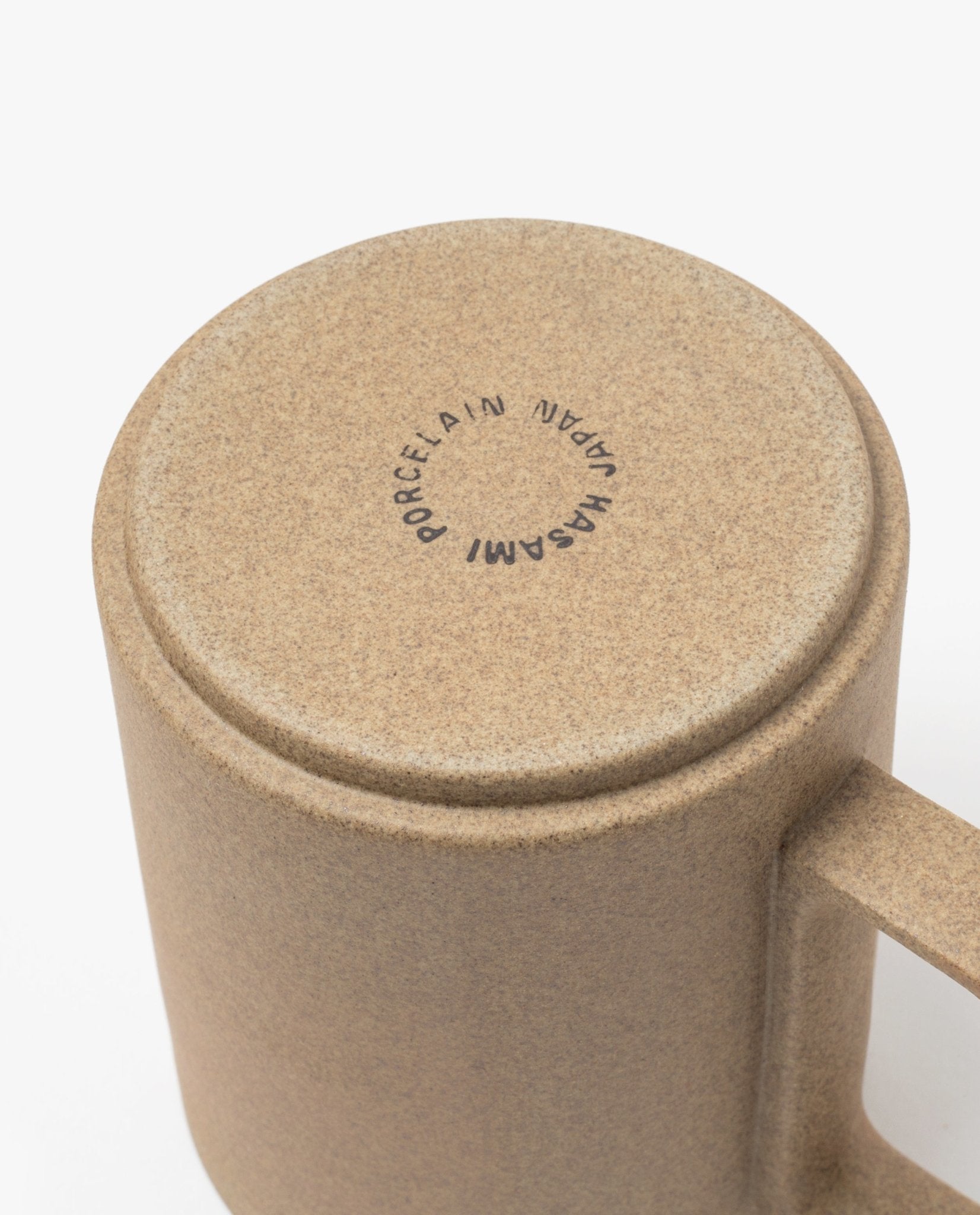 A close-up of the base of the Hasami Porcelain Mug (Natural) showcases its beige, speckled handmade texture. The white bottom is stamped in a circular pattern with "HASAMI PORCELAIN JAPAN," emphasizing the craftsmanship and artistry of Japanese ceramics.