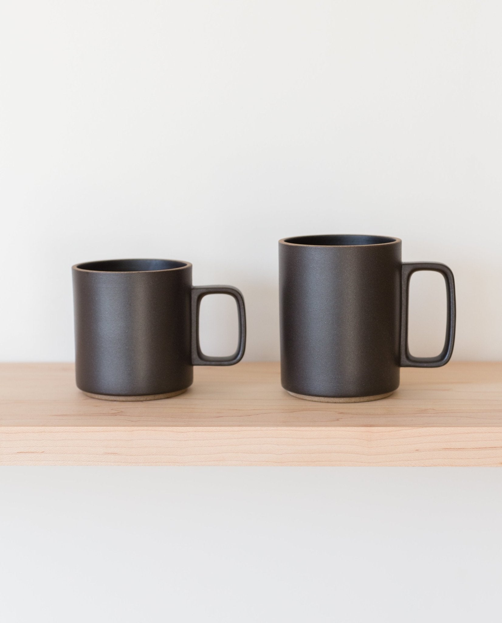 Two Hasami Porcelain Mugs (Matte Black) with square handles sit side by side on a light wooden shelf against a plain white background, one slightly shorter and wider than the other, showcasing subtle handmade charm.