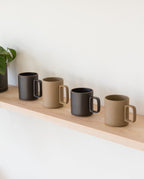 Four Hasami Porcelain Mugs (Matte Black), inspired by handmade Japanese design, alternate with beige mugs on a light wooden shelf against a white wall. A black-potted plant partially appears on the shelf’s left side.