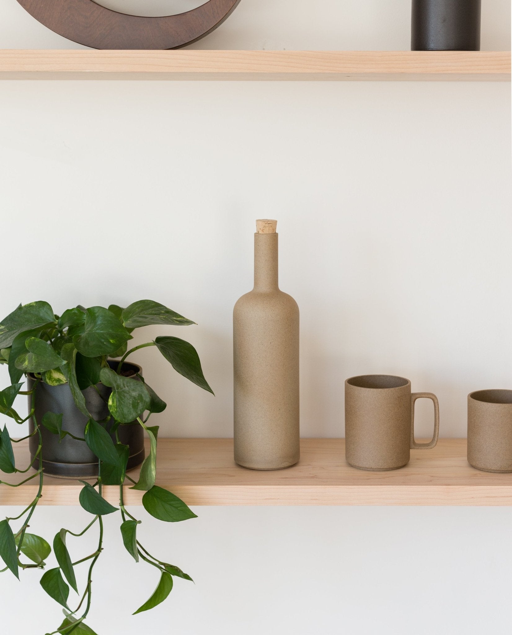 A wooden shelf displays a green potted plant, a tall ceramic bottle with a cork, and two brown Japanese ceramic mugs—including the Hasami Porcelain Mug (Natural)—arranged elegantly against a white wall.