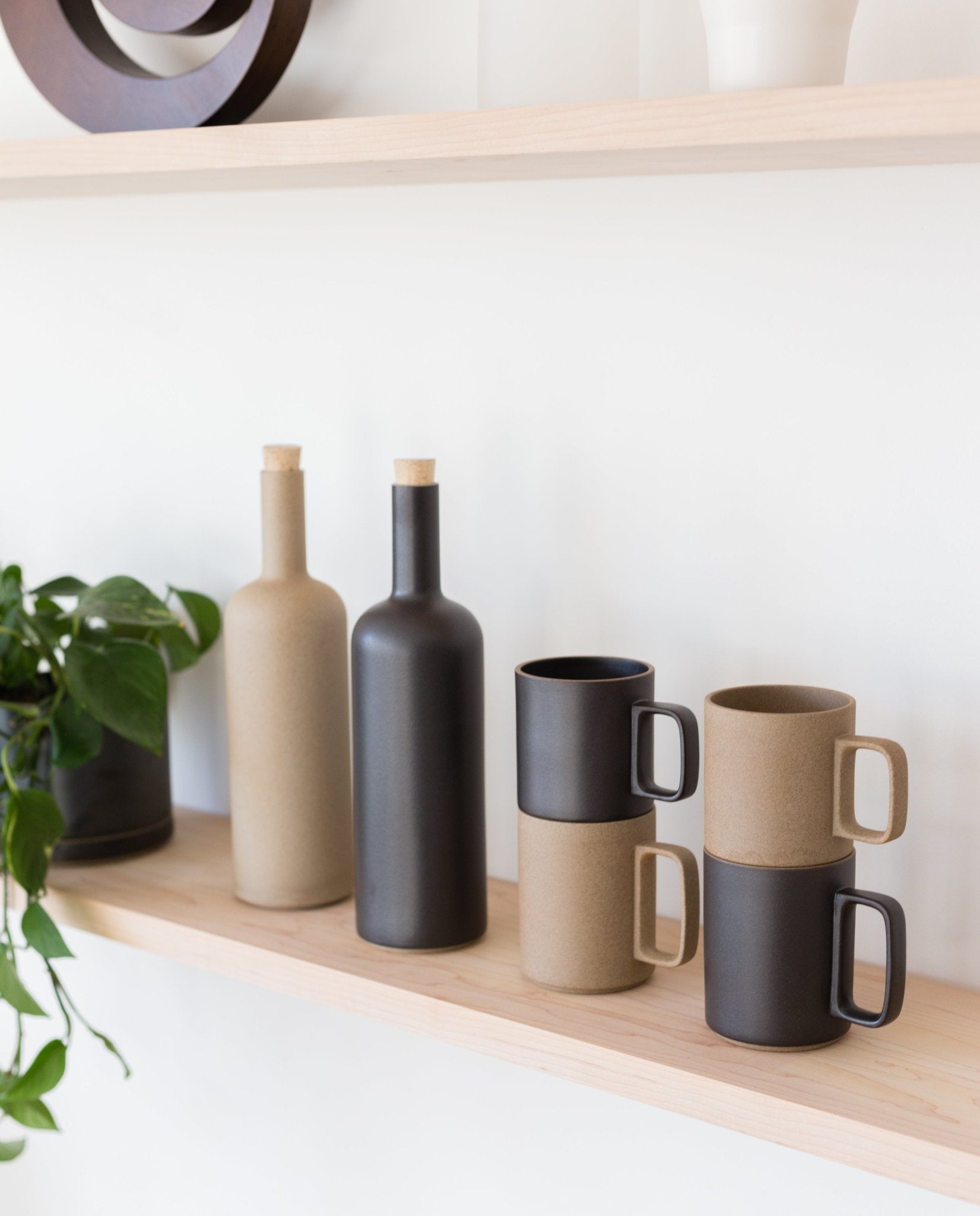 Two Hasami Porcelain Mugs (Matte Black) are stacked on a light wooden shelf beside two bottles with cork stoppers and a leafy green potted plant, all set against a white wall.