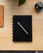 A black spiral-bound notebook with the INVENTERY Bolt Action Pen (Stainless) placed diagonally on top sits on a light wooden desk. A portion of a tan leather item, a plant leaf, and a glass hourglass are also visible.