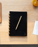 An INVENTERY Bolt Action Pen (Brass) rests on a closed black notebook with gold rings, placed on a light wooden desk. Nearby, there is a small notepad and part of a yellow object visible.