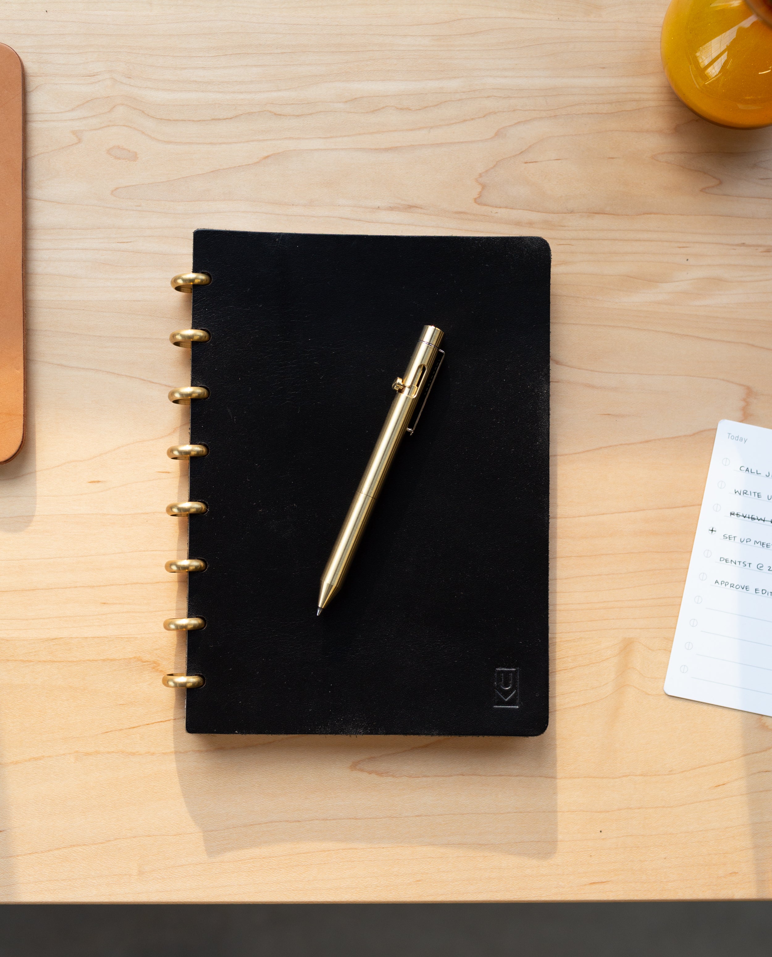 An INVENTERY Bolt Action Pen (Brass) rests on a closed black notebook with gold rings, placed on a light wooden desk. Nearby, there is a small notepad and part of a yellow object visible.