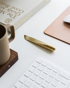 A Craighill Desk Knife (Brass) rests on a white desk beside a keyboard, brown mug, mouse pad, and a book—a stylish multipurpose cutting tool for your workspace.