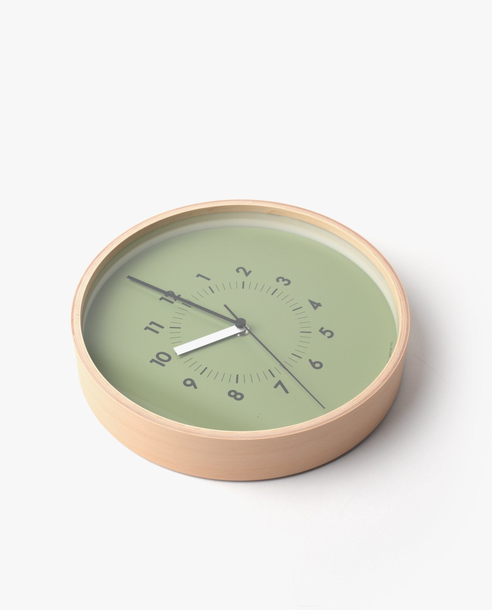 The Lemnos SOSO Clock (Green) features a round plywood design with a light wood frame and a green face. Black numbers spiral across the surface, complemented by black hour and minute hands. Resting elegantly on a white surface, this minimalist clock is an ideal addition to any modern space.