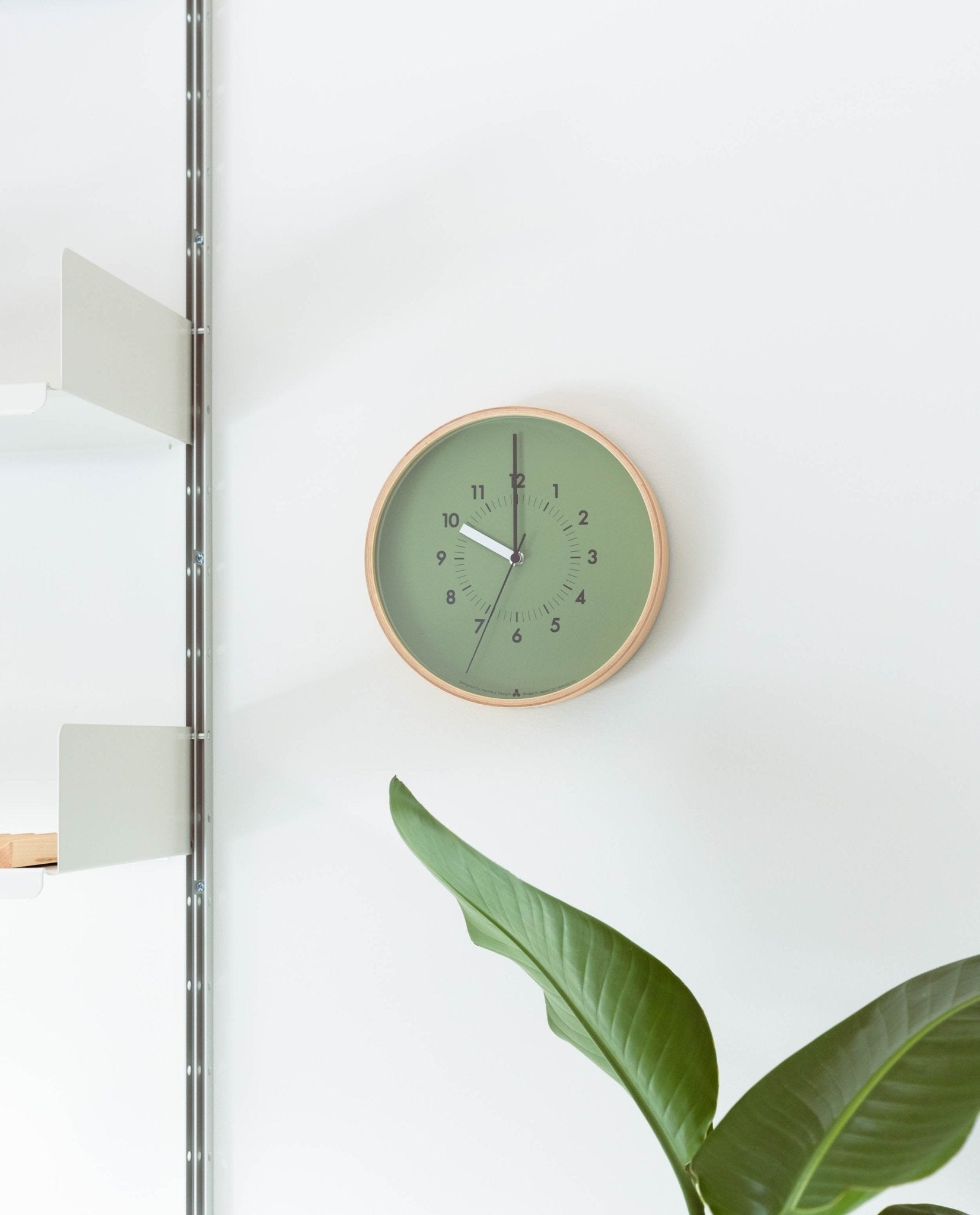 A Lemnos SOSO Clock (Green) displays the time as 12:03. A leafy plant is visible in the lower right corner, and white shelves are mounted on the left side of the white wall.