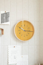 A Lemnos SOSO Clock (Orange) hangs on a tiled wall above pinned papers—among them a "VICTORY" sign and sheets with sketches and notes—its silent sweeping second hand complementing the calm atmosphere of this creative or office workspace. The minimalist round design displays 10:10.