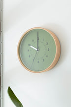 The Lemnos SOSO Clock (Green), a round minimalist wall clock with a light plywood frame and green face, shows the time as 11:00:49. This silent clock is mounted on a white wall near a green plant leaf.