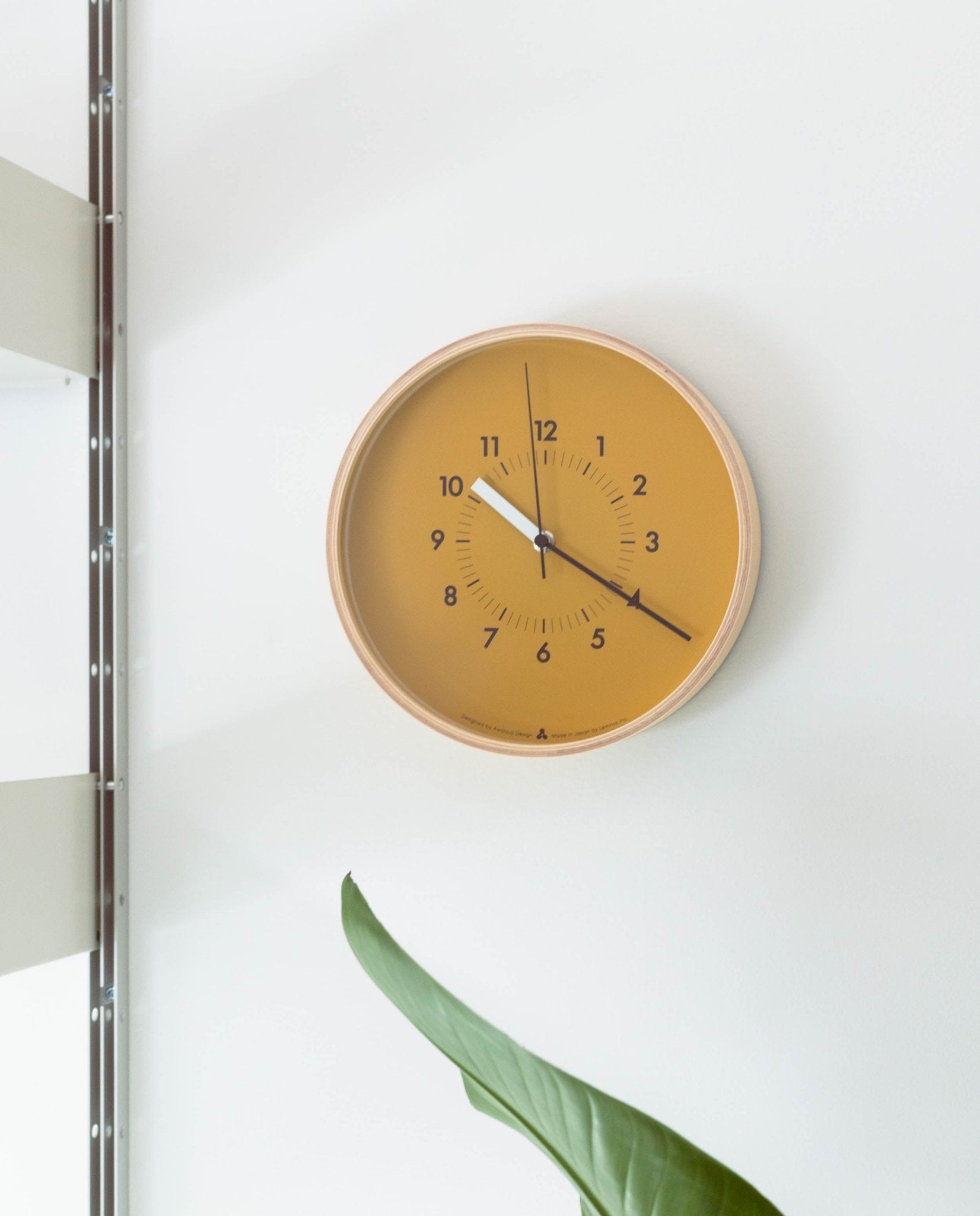 A Lemnos SOSO Clock (Orange) shows the time as 11:58. The round minimalist clock is mounted on a white wall near a shelf, with a green plant partially visible at the bottom.