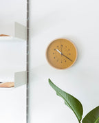 A Lemnos SOSO Clock (Orange) displays the time as 11:05. A green leafy plant sits in the lower right corner, while white shelves appear on the left against a white wall.