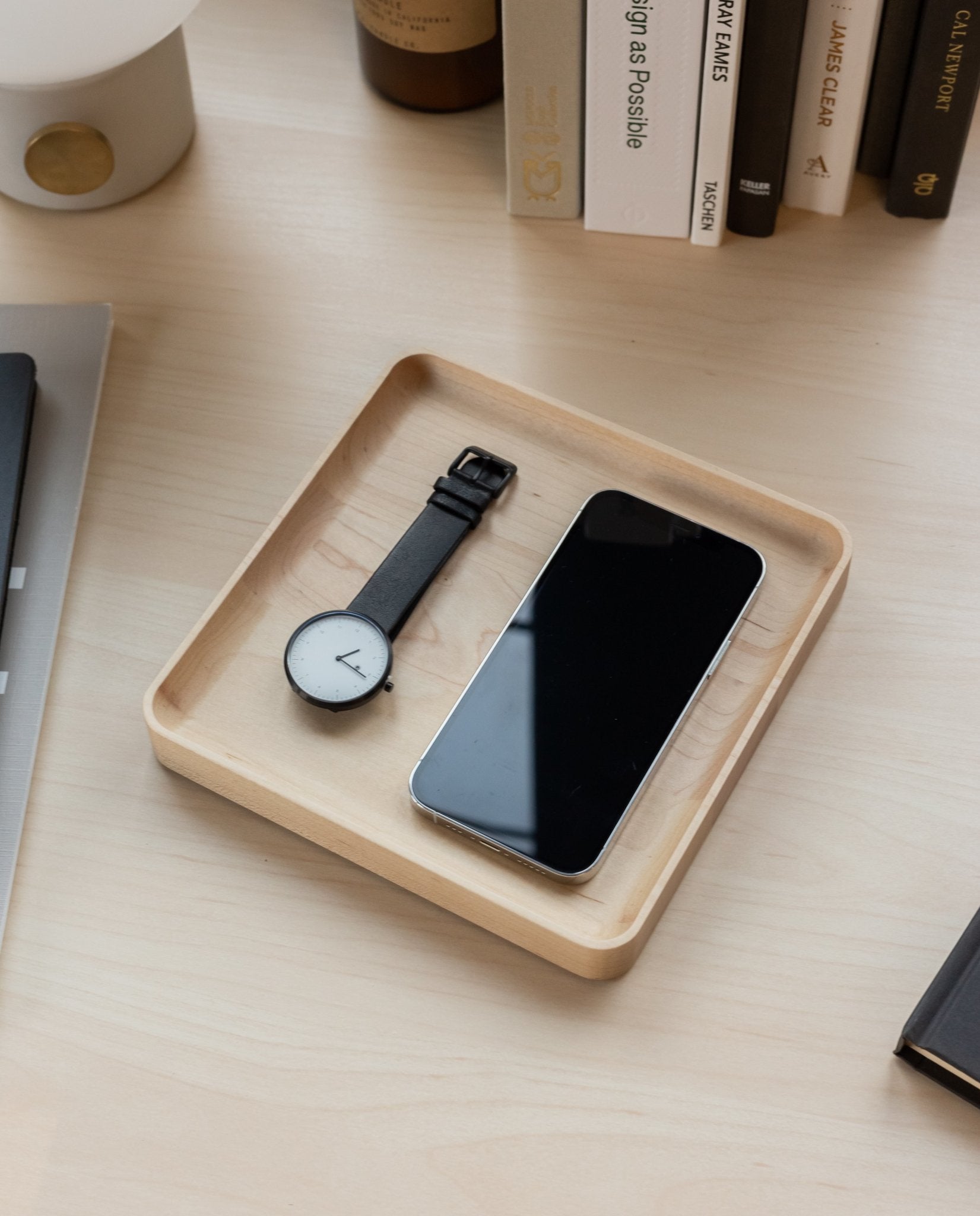 A black wristwatch and a smartphone sit side by side in a handcrafted Valet Tray (Maple - Medium) on a tidy desk, with books and other items visible in the background.