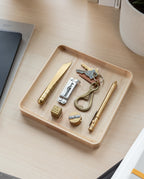 A Valet Tray (Maple - Medium) sits on a light desk, holding brass items—a pen, knife, multi-tool, keychain with keys, dice, a round object, and a small cylinder. Nearby are a plant pot and some notebooks.