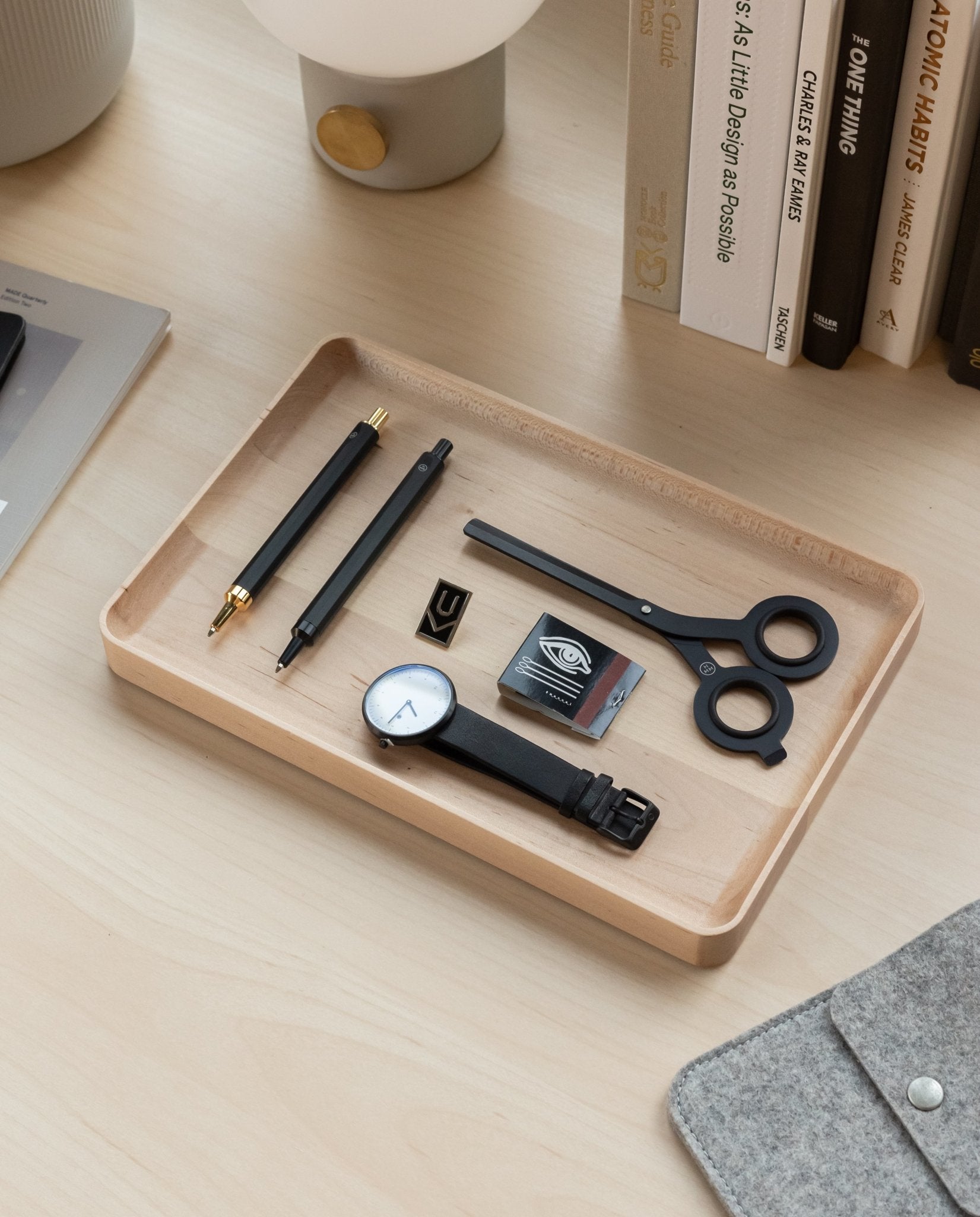 The Valet Tray (Maple - Large) sits on a desk holding two pens, scissors, a black wristwatch, an eraser, a pencil sharpener, and a small block, with books and a grey felt item nearby.