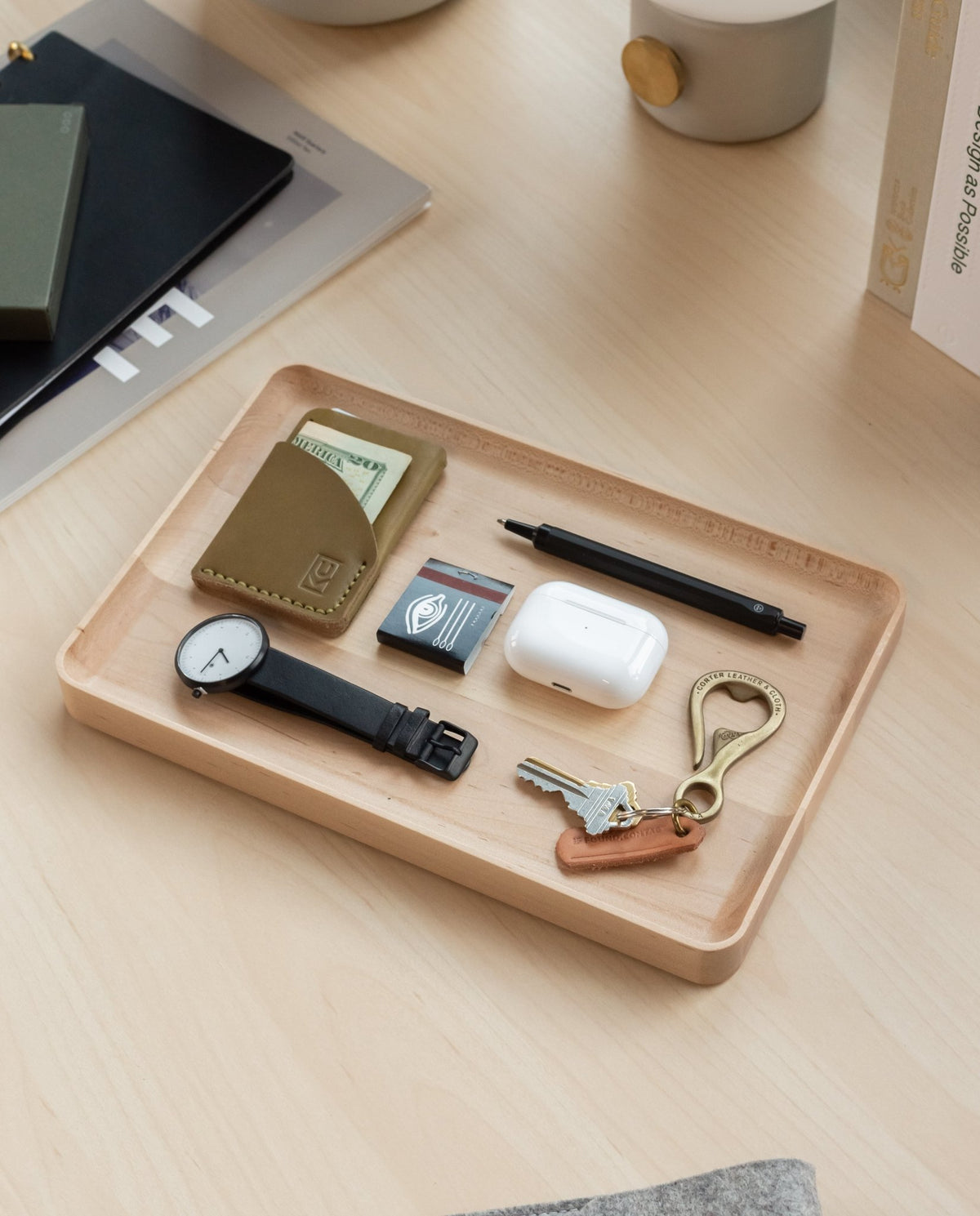 The Valet Tray (Maple - Large) neatly holds a wallet, cash, a watch, a lighter, a pen, wireless earbuds, a brass key hook, and a set of keys on a light wood desk.