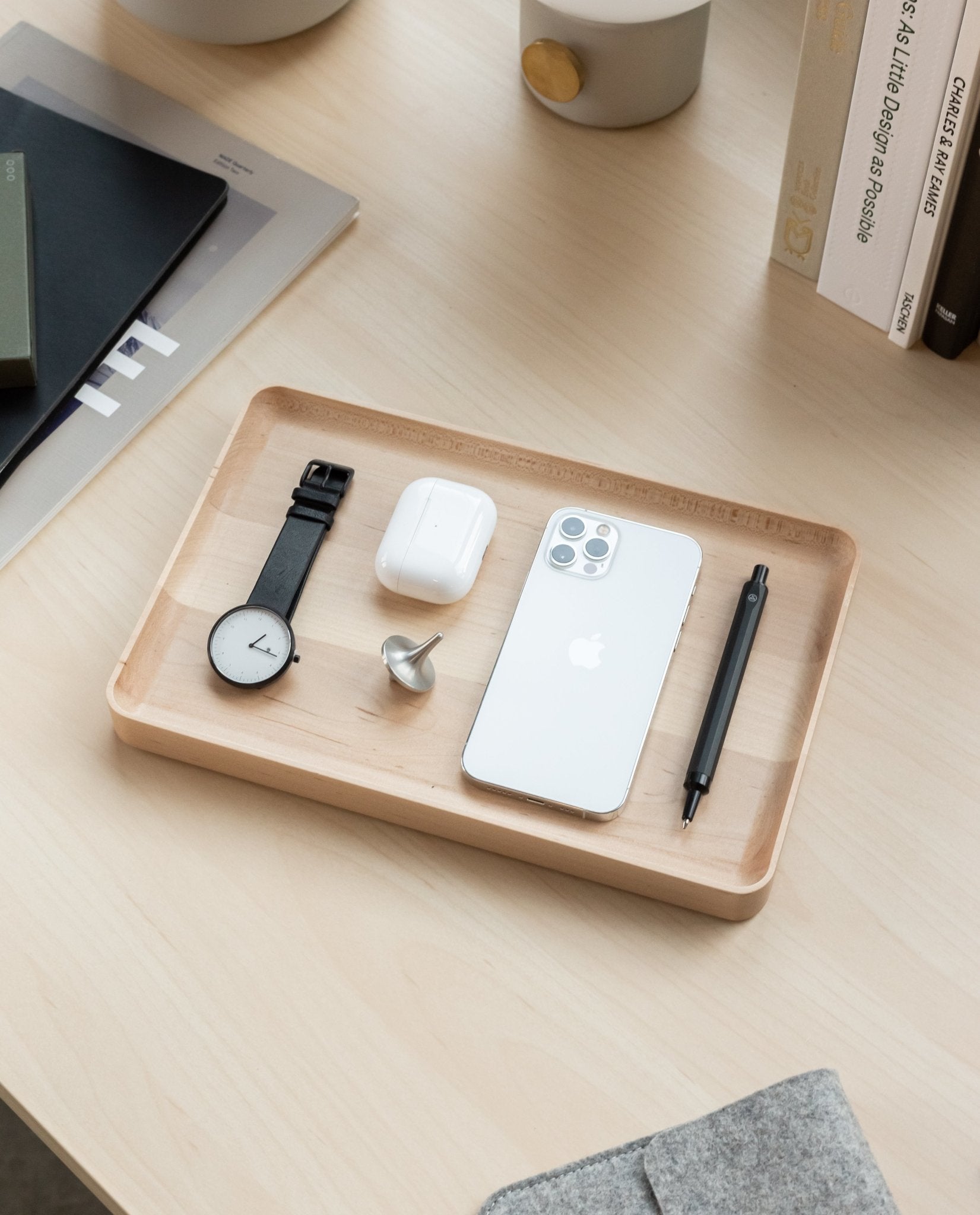 On a light desk, the Valet Tray (Maple - Large) holds a black watch, a white wireless earbuds case, a silver spinning top, a white smartphone, and a black pen; nearby are books and a lamp.