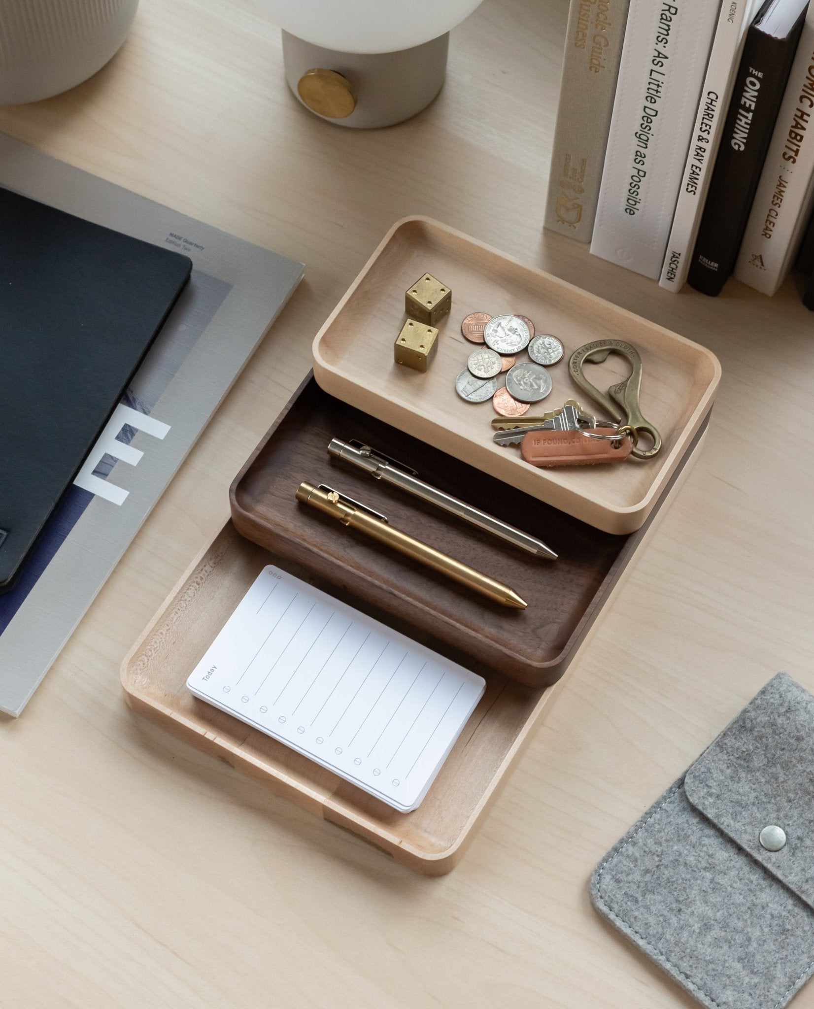 Three handcrafted Valet Trays (Maple - Small) neatly hold pens, a notepad, coins, keys, dice, and a USB drive on an organized desk beside books, a lamp, and a felt pouch. Each valet tray is crafted from solid maple for enduring elegance and organization.