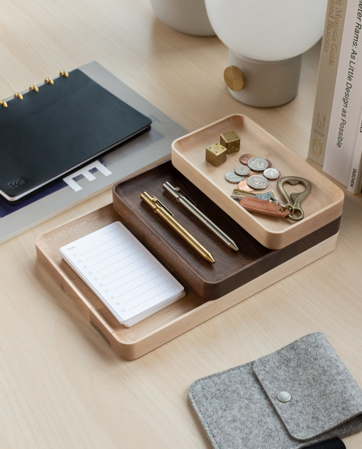 A Valet Tray (Maple - Large) sits on the desk, neatly holding pens, coins, keys, and a notepad. Nearby are a notebook, a grey felt pouch, a lamp, and stacked books for an organized workspace.