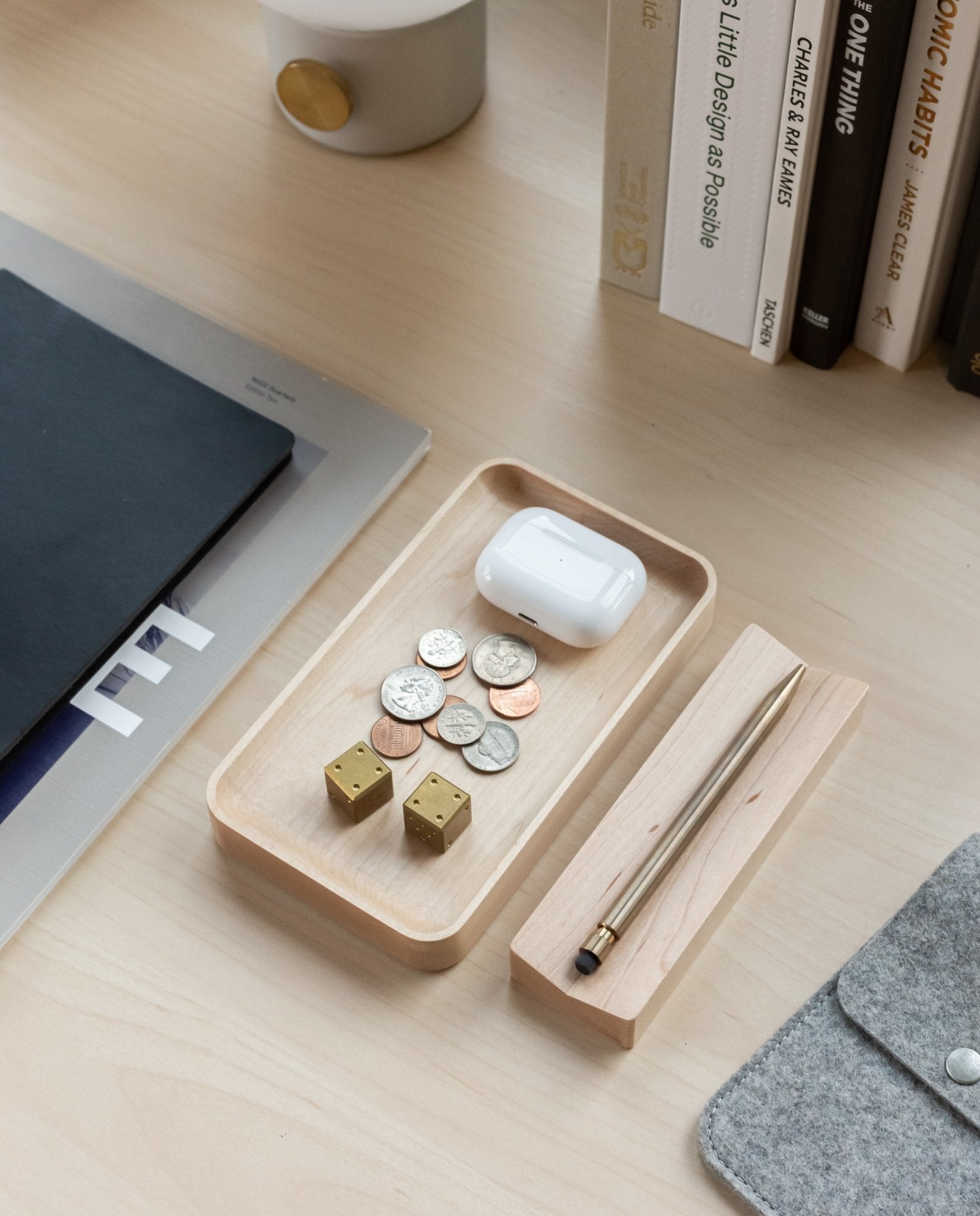 A Valet Tray (Maple - Small) sits on a desk, holding coins, two gold dice, and wireless earbuds. Nearby, a pen rests on a wooden holder, with books and folders arranged in the background.