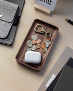 The Valet Tray (Walnut - Small), handcrafted in Pennsylvania, sits on a desk holding coins, a brass keychain bottle opener, two keys, and a white wireless earbud case beside a pen, notebooks, and a gray felt pouch for everyday carry.