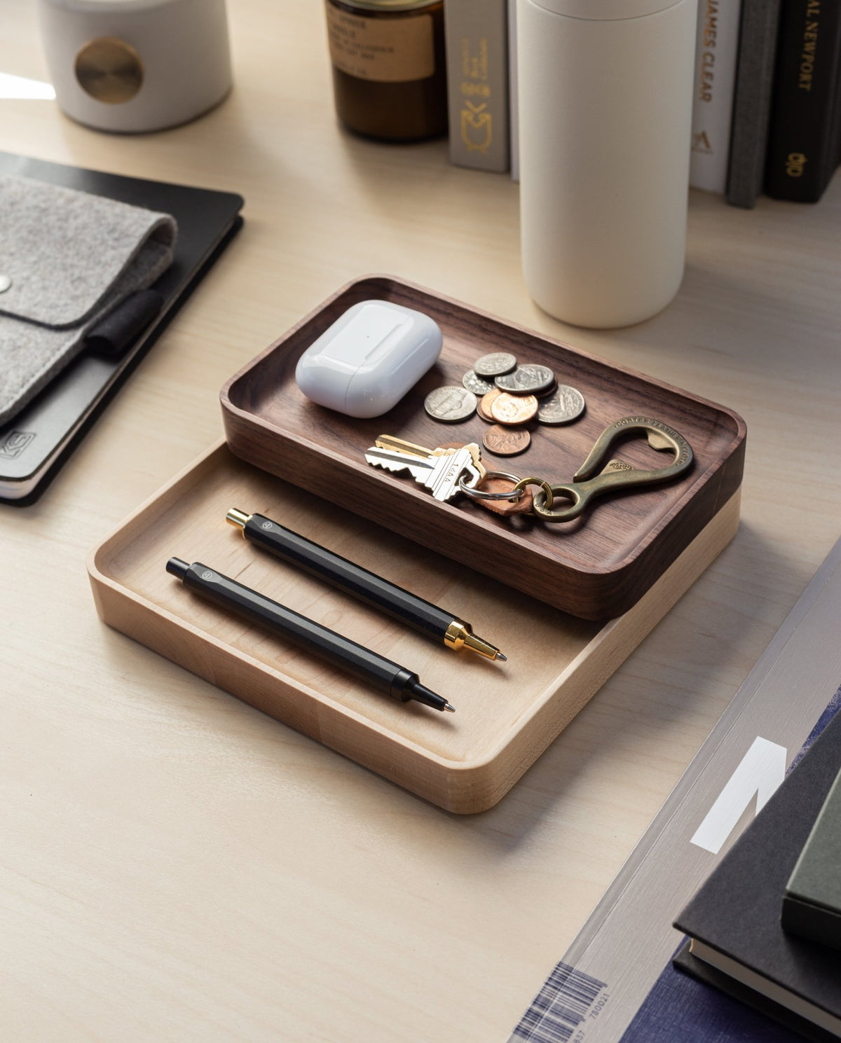 Two handcrafted Valet Tray (Maple - Medium) trays on a desk neatly hold pens, keys, coins, a keychain, and wireless earbuds. Nearby are a felt pouch, white bottle, and books, creating an organized workspace atmosphere.