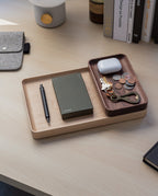 The Valet Tray (Walnut - Small), handcrafted in Pennsylvania, keeps your pen, green notebook, bottle opener, keys, coins, and white wireless earbuds organized on your desk—ideal for everyday carry storage near books and a gray felt pouch.