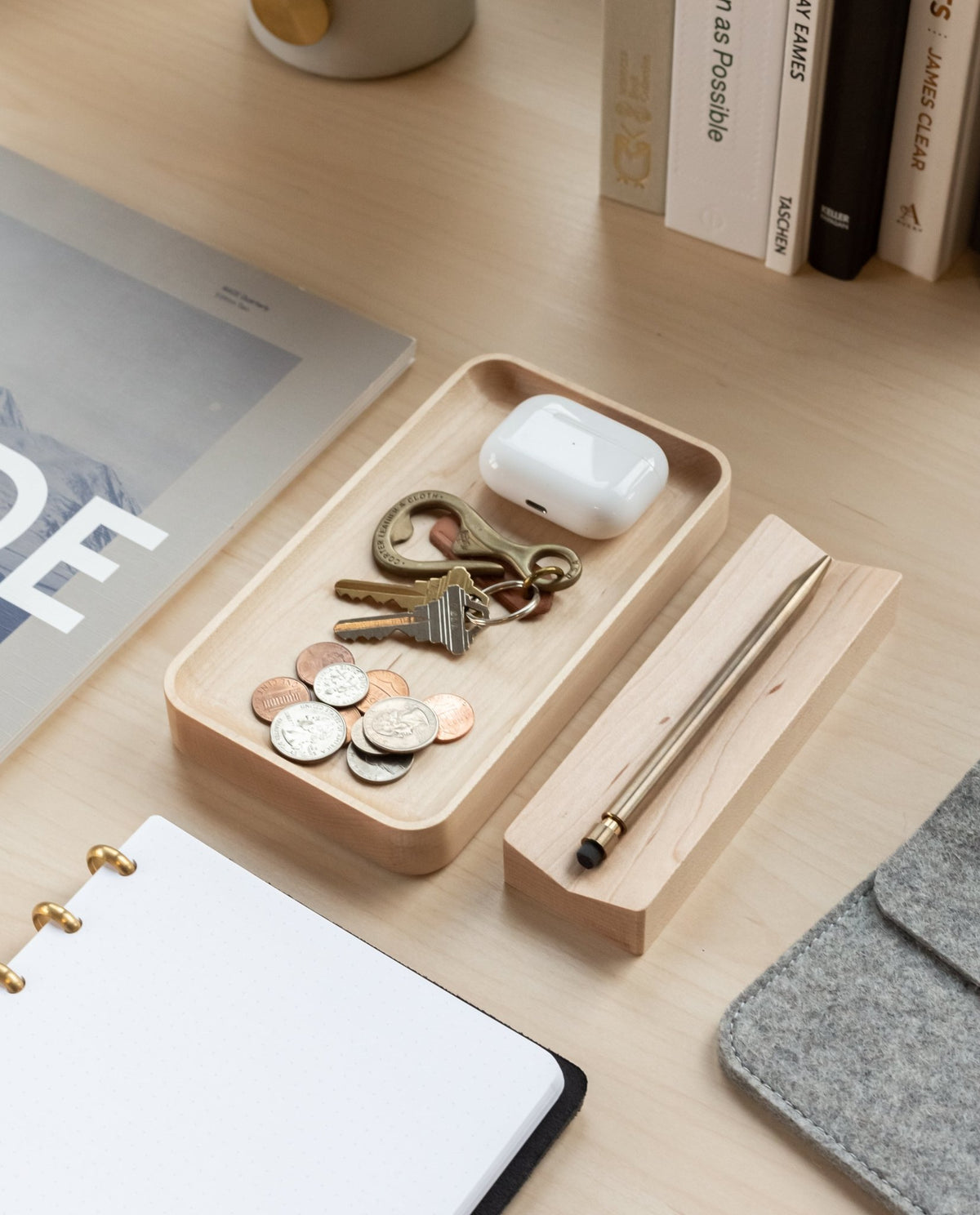 A Valet Tray (Maple – Small), handcrafted from solid maple, holds coins, keys, and wireless earbuds on a light desk. Nearby are a stylus in its holder, an open notebook, a magazine, several books, and part of a laptop sleeve.