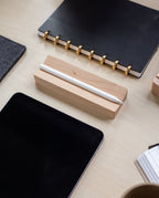 A Pen Tray (Maple), made in Pennsylvania, holds a stylus on a desk beside a tablet, a black notebook with gold rings, and a cup, all neatly arranged on a light wood surface.