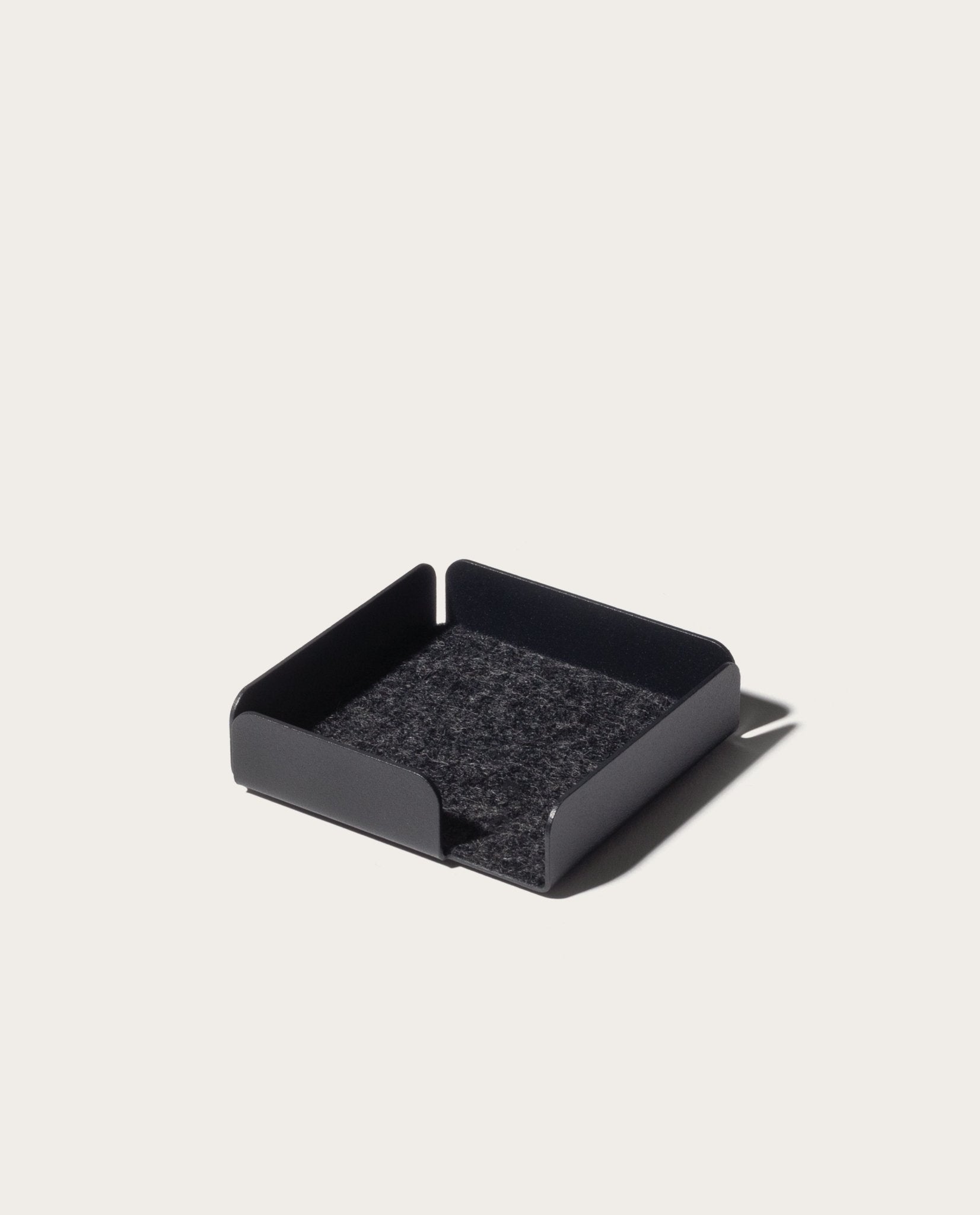 The Gather Note Tray (1x1 - Black) is a sleek black square tray with short sides and a soft, dark felt bottom. Placed against a light background, this elegant Magnetic Desk Organizer helps keep your workspace tidy and clutter-free.