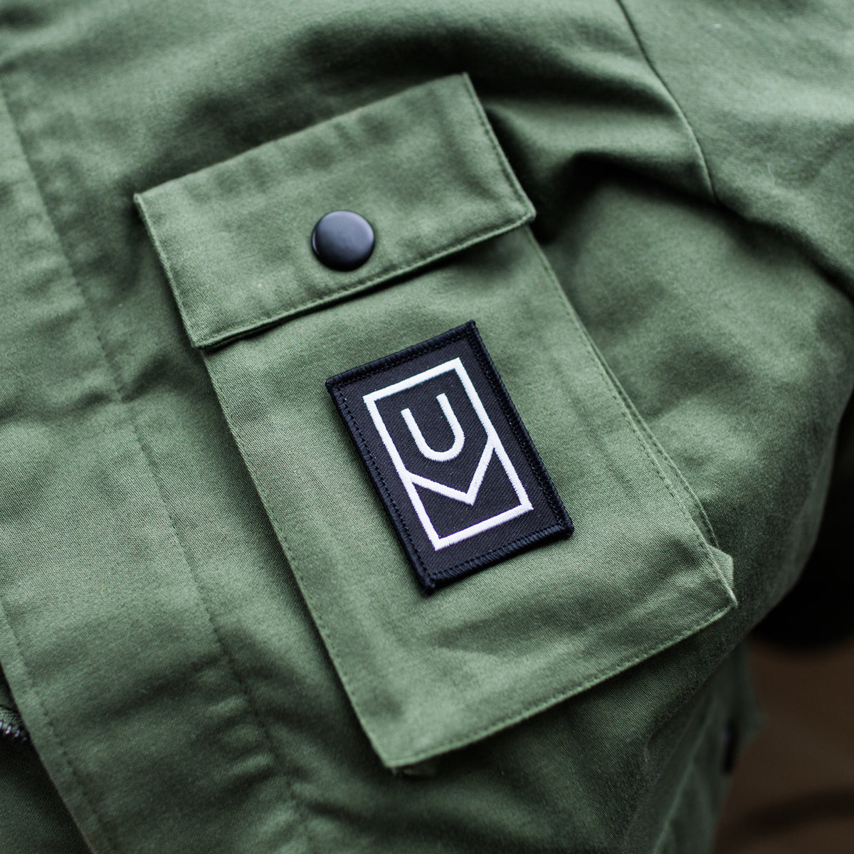 A close-up of an olive green jacket pocket with the Ugmonk Patch, a black and white rectangular embroidered patch featuring a geometric design, stitched below a buttoned flap.