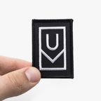A hand holds the Ugmonk Patch, a rectangular black patch featuring bold white geometric lines that form abstract shapes—possibly suggesting the letters U and V—against a plain white background.